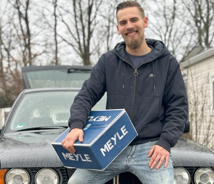Janik Lindemann, Geschäftsführer von KFZ-Teile Lindemann, mit einem Ersatzteilkarton vor einem Fahrzeug